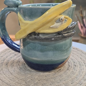 May include: A handmade ceramic mug with a unique design. The mug is primarily blue with a yellow sculpted banana and a small white bird. The handle is a loop with a small blue accent. The mug sits on a woven placemat.