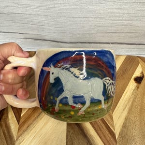 Może przedstawiać: Ceramiczny kubek z błyszczącym wykończeniem, przedstawiający ręcznie malowanego białego jednorożca z różowym rogiem. Tło ukazuje ciemnoniebieskie niebo z żywym tęczowym łukiem nad zielonym polem usianym małymi, kolorowymi akcentami kwiatowymi.