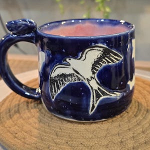 May include: A ceramic mug with a dark blue exterior and a pink interior. The mug features a white and black illustration of a bird in flight. The handle has a decorative flourish.