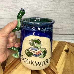 May include: A handmade ceramic mug with a green, snake-shaped handle. The mug is blue and cream-coloured, with a cartoon illustration of a green bookworm and the word "BOOKWORM" on the front. The mug is held by a hand.