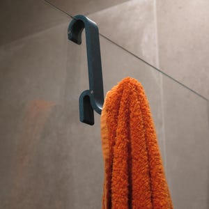 May include: A dark teal, speckled S-shaped shower hook hanging over the top edge of a glass panel. The hook features a curved top section for mounting and a lower hook section that holds a bright orange, textured cotton bath towel.