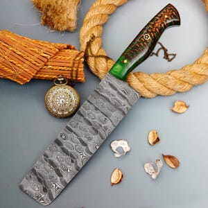 May include: A Damascus steel chef's knife with a patterned blade and a green and brown handle. The knife is displayed with a rope, a pocket watch, and decorative elements. The blade has a unique, wavy pattern.