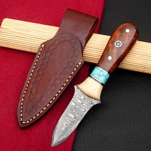 May include: A handcrafted knife with a patterned steel blade, wooden handle, and turquoise accents. The knife is paired with a brown leather sheath. The handle features decorative inlays. The knife is resting on a bamboo mat and red fabric.