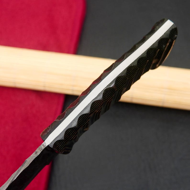 May include: Close-up of a knife with a textured, dark handle and a white stripe down the center. The blade has a patterned design. The knife is set against a red and black background.