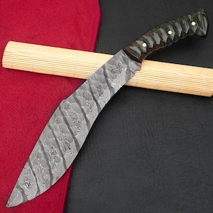 May include: A curved, Damascus steel knife with a dark green and black handle. The blade has a unique pattern and is resting on a bamboo mat and a red cloth.