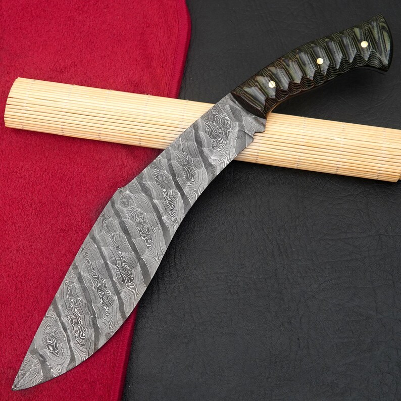 May include: A curved, Damascus steel knife with a dark green and black handle. The blade has a unique pattern and is resting on a bamboo mat and a red cloth.