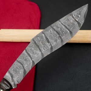 May include: Close-up of a Damascus steel knife blade with a unique, swirling pattern. The blade has a curved shape and a sharp edge. The handle is not visible. The knife rests on a wooden dowel and a red fabric.