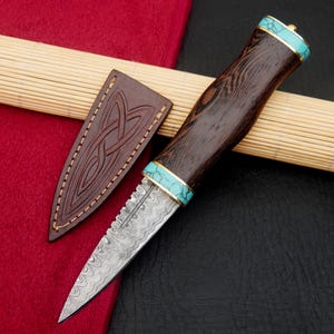 May include: A handcrafted knife with a patterned steel blade and a dark wood handle. The handle is accented with turquoise and gold-colored bands. Includes a brown leather sheath with an embossed Celtic knot design.