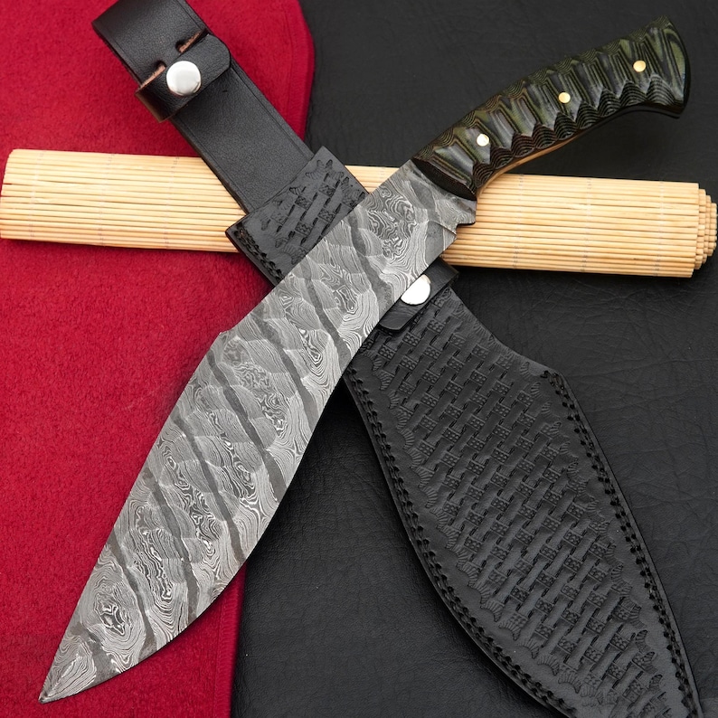 May include: A Damascus steel knife with a patterned blade and a green and black handle, resting in a black leather sheath. The sheath has a woven texture. A leather strap secures the knife. The knife is on a red and black surface.