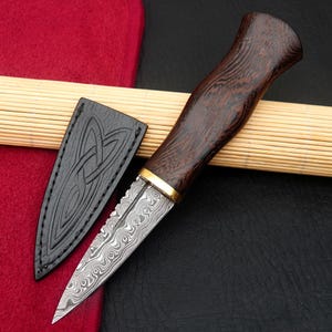May include: A handcrafted dagger with a patterned steel blade and a dark wood handle. The handle is accented with a gold-colored band. Includes a black leather sheath with an embossed Celtic knot design.