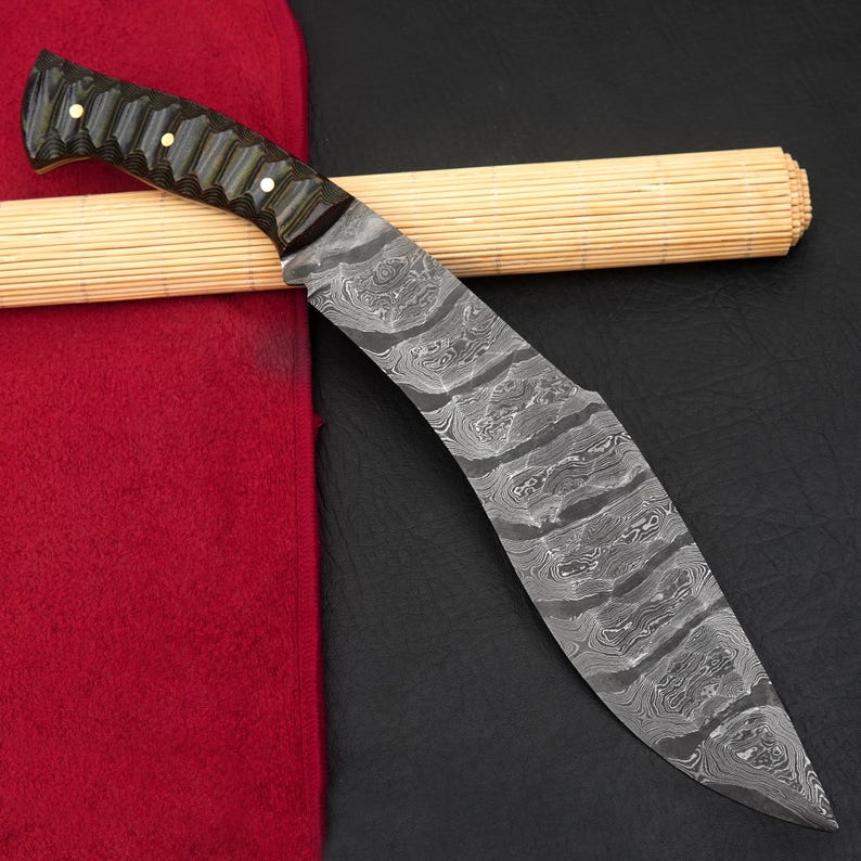 May include: A Damascus steel knife with a curved blade and a dark wooden handle. The handle is accented with small, gold-colored rivets. The knife rests on a red cloth and a bamboo mat.