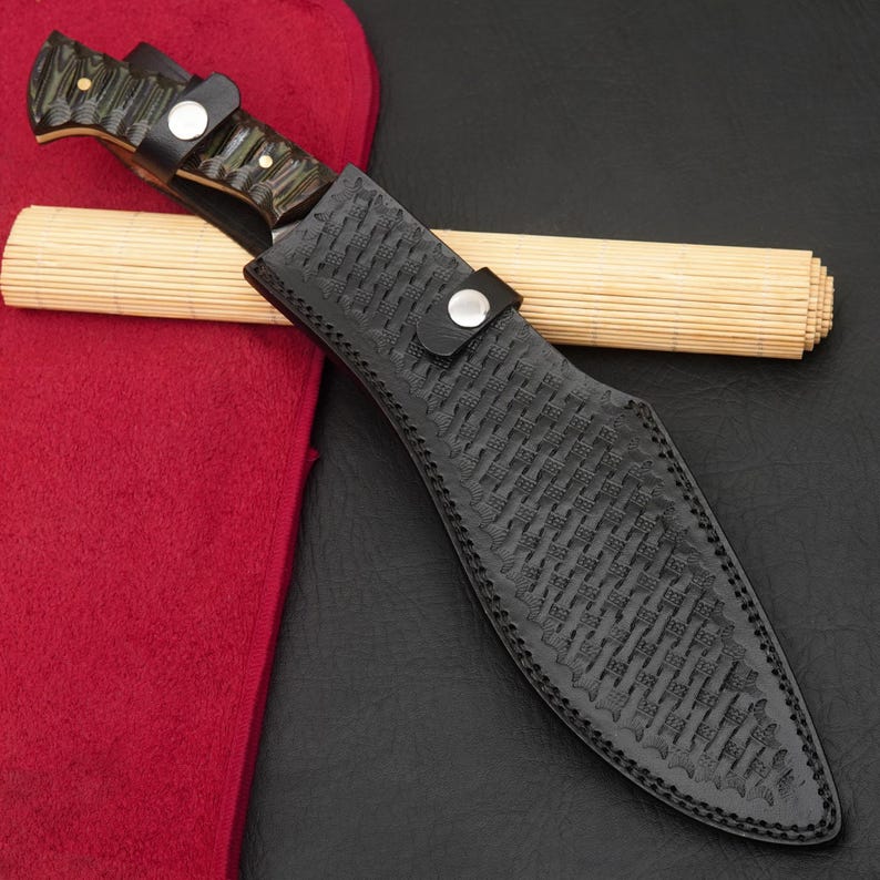 May include: A handcrafted knife with a dark, patterned handle and a black leather sheath. The sheath has a woven texture and a strap with a silver button. A rolled bamboo mat rests behind the knife.