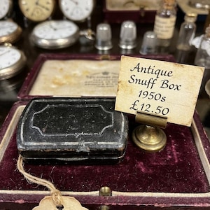 May include: A rectangular black snuff box with a decorative border, resting in a deep purple velvet-lined case. A small sign on a brass stand displays the text: Antique Snuff Box 1950s £12.50. A beige paper tag is attached to the box with twine.