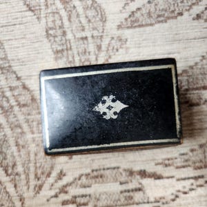 May include: A rectangular black box with a smooth, dark finish. The lid features a thin, light-coloured border framing the edges and a centred, intricate, light-coloured decorative emblem with a four-pointed, stylised floral design.
