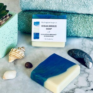 May include: A bar of handmade soap with a blue, green and white design. The soap is labelled "Ocean Breeze Soap" with the text "The Fragrant Soap Co." and "with Eucalyptus, Bergamot & Cedarwood".