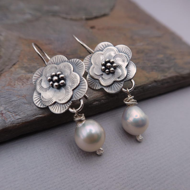 May include: A pair of silver-toned flower earrings with a pearl drop. The earrings feature detailed floral designs with a cluster of small beads at the center of each flower. The pear-shaped pearls are attached with wire wrapping.