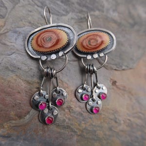 May include: A pair of silver earrings with oval-shaped, wood-like center stones. The earrings have a dangling design with three small, silver discs, each holding a pink gemstone. The earrings are designed with a rustic, bohemian style.