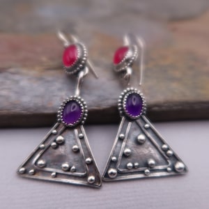 May include: A pair of sterling silver drop earrings featuring a triangular base with a textured, oxidized finish and small raised silver dots. Each earring is accented with a small oval purple gemstone at the top of the triangle and a smaller pink oval gemstone set above it on the hook.