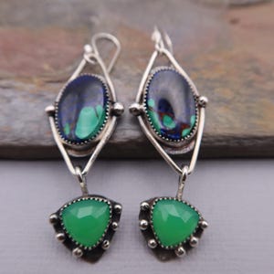 May include: A pair of silver earrings with a geometric design. Each earring has an oval blue and green stone at the top and a triangular green stone at the bottom. The stones are set in a silver frame with decorative accents.