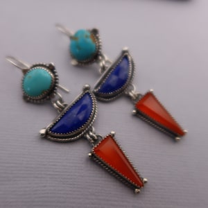 May include: A pair of silver earrings with turquoise, blue lapis lazuli, and red carnelian gemstones. The earrings feature a turquoise stone at the top, a half-moon of blue lapis, and a red carnelian triangle pendant. The jewelry has a Southwestern style.