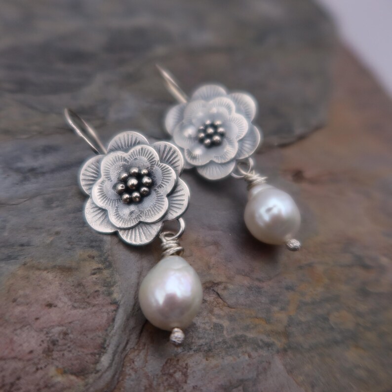 May include: Silver flower earrings with a pearl drop. The earrings feature detailed, openwork flower designs with a cluster of small beads at the center. Each earring has a pear-shaped, white pearl suspended below the flower.
