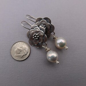 May include: Silver flower earrings with a pearl drop. The earrings feature detailed floral designs with a small pearl at the center of each flower. The earrings have a hook closure and a large, white, teardrop-shaped pearl.