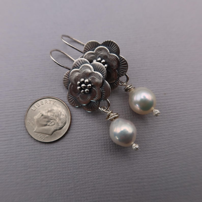 May include: Silver flower earrings with a pearl drop. The earrings feature detailed floral designs with a small pearl at the center of each flower. The earrings have a hook closure and a large, white, teardrop-shaped pearl.