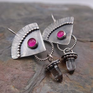 Puede incluir: Pendientes de plata con un diseño en forma de abanico, con una gema rosa en el centro y un borde con cuentas. Cada pendiente tiene un lazo de plata retorcido y un colgante de cristal transparente con una tapa de plata.