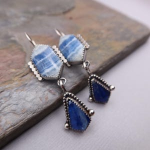 May include: A pair of silver earrings with blue and white gemstone pendants. The top portion of each earring features a light blue and white stone in a geometric shape, while the bottom pendant is a deep blue, teardrop-shaped stone.