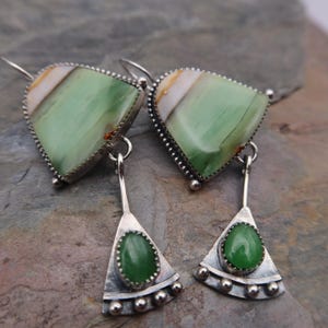 Pode incluir: Um par de brincos pendentes com uma pedra grande triangular de faixas verde-pálido e branco engastada em prata, ligada por uma fina barra de prata a uma base triangular menor com uma pedra lisa de cor verde-escura.