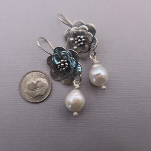 May include: A pair of silver-toned flower earrings with a pearl drop. The earrings feature detailed floral designs with a cluster of small beads at the center of each flower. The pearls are white and teardrop-shaped.
