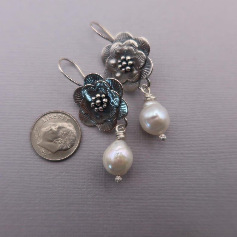 May include: A pair of silver-toned flower earrings with a pearl drop. The earrings feature detailed floral designs with a cluster of small beads at the center of each flower. The pearls are white and teardrop-shaped.
