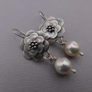 May include: Silver flower earrings with pearl drops. The earrings feature detailed, textured flower designs with a cluster of small beads at the center. Each earring has a large, lustrous, white pearl suspended below the flower.