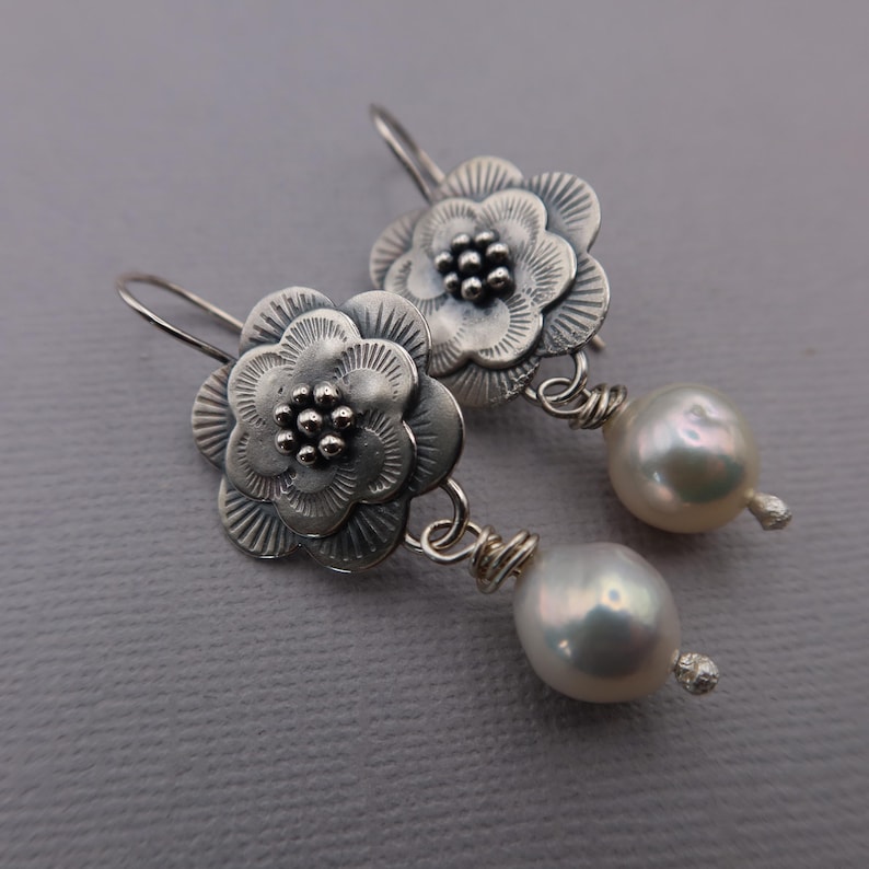 May include: Silver flower earrings with pearl drops. The earrings feature detailed, textured flower designs with a cluster of small beads at the center. Each earring has a large, lustrous, white pearl suspended below the flower.