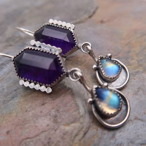 May include: A pair of drop earrings featuring a faceted hexagonal purple amethyst stone set in silver with a decorative scalloped edge. Below each amethyst hangs a smaller, teardrop-shaped iridescent moonstone set in a circular silver frame.