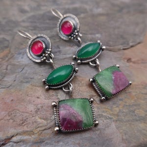 May include: A pair of sterling silver drop earrings featuring a circular top with a round pink stone, a central marquise-shaped green stone, and a square bottom stone with a mottled green and pink pattern, all set in decorative metal frames.