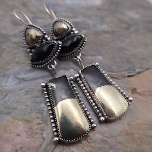 Puede incluir: Pendientes de plata con diseño geométrico. Cada pendiente presenta una combinación de elementos negros, dorados y plateados. Los pendientes tienen un diseño en capas con una mezcla de formas, incluyendo triángulos y rectángulos.