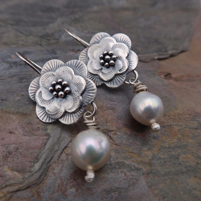 May include: A pair of silver flower earrings with a pearl drop. The earrings feature detailed flower petals and a cluster of small beads at the center. The pearls are a soft white color and are suspended from the flower.