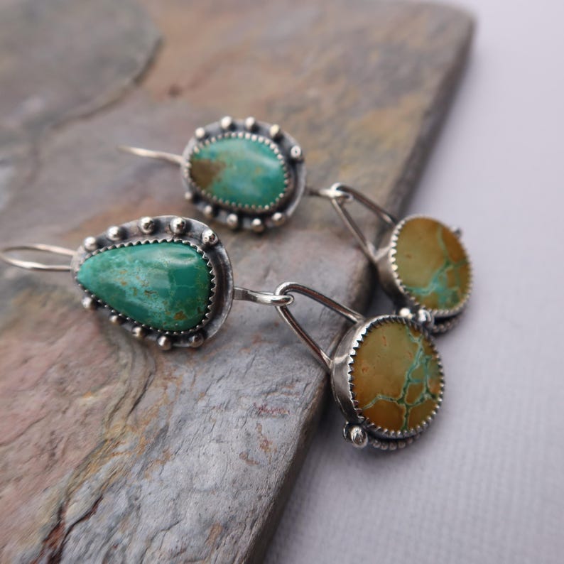 May include: Silver earrings with turquoise and tan-colored stones. The earrings feature a teardrop-shaped turquoise stone and a round turquoise stone, both set in a silver bezel with a beaded edge. The earrings are connected by silver links.