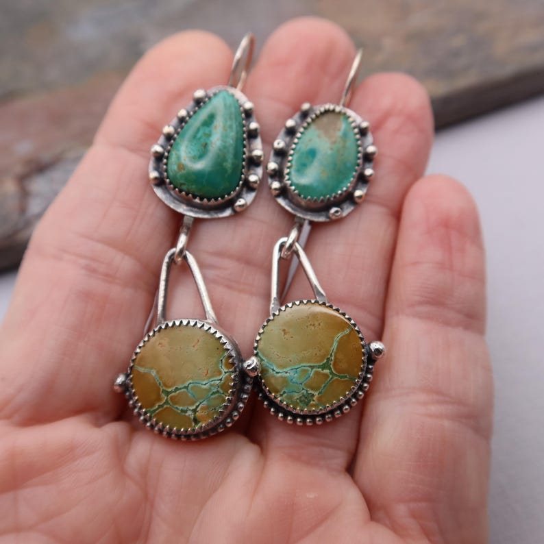 May include: A pair of silver earrings with turquoise and tan-colored stones. The earrings have a teardrop-shaped turquoise stone at the top, connected to a circular tan stone with turquoise veining. The stones are set in a silver bezel with a beaded edge.