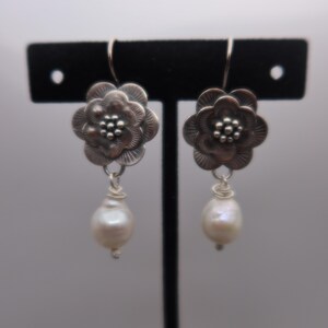 May include: A pair of silver-toned flower earrings with a pearl drop. The earrings feature a detailed floral design with a small cluster of beads at the center. The teardrop-shaped pearls are white and dangle from the flower.
