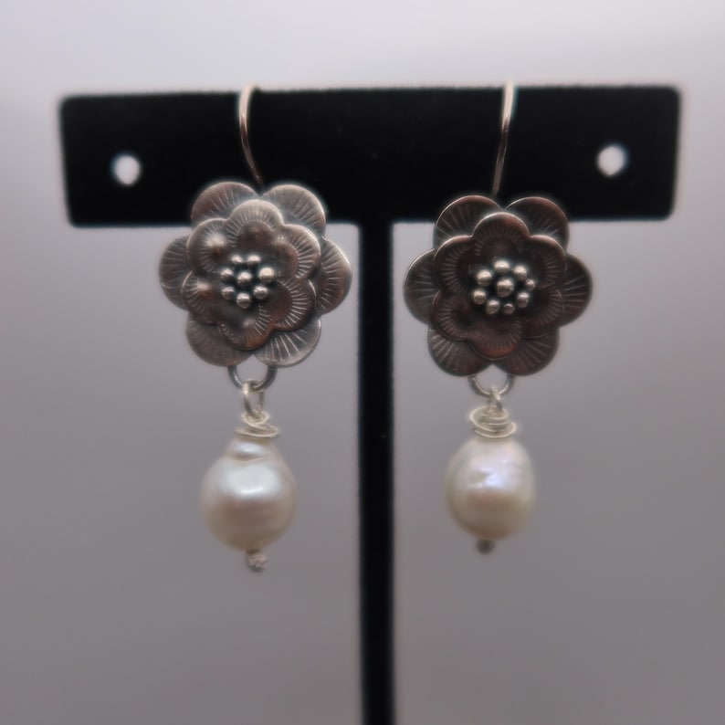 May include: A pair of silver-toned flower earrings with a pearl drop. The earrings feature a detailed floral design with a small cluster of beads at the center. The teardrop-shaped pearls are white and dangle from the flower.