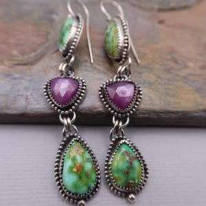 May include: A pair of dangle earrings with a silver finish. Each earring features three gemstones: a green oval, a pink heart, and a green oval. The gemstones are set in a beaded silver frame. The earrings have a hook closure.