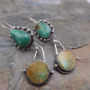 May include: Silver earrings with turquoise and tan-colored stones. The earrings have a detailed, textured silver setting with a teardrop-shaped turquoise stone and a round tan stone with turquoise veining. The earrings are displayed on a gray stone surface.