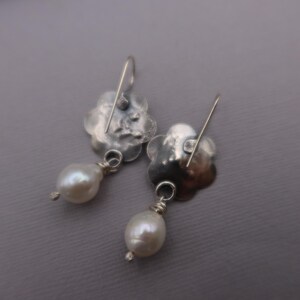 May include: A pair of silver-toned earrings. Each earring features a flower-shaped top with a dangling, pear-shaped white pearl. The earrings have a hook closure. The background is a solid gray.