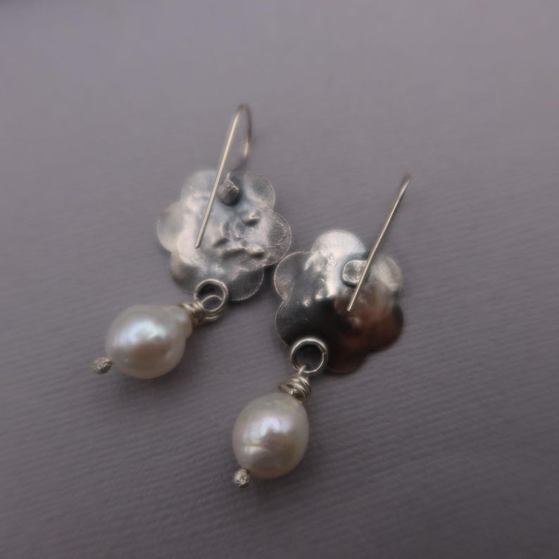 May include: A pair of silver-toned earrings. Each earring features a flower-shaped top with a dangling, pear-shaped white pearl. The earrings have a hook closure. The background is a solid gray.