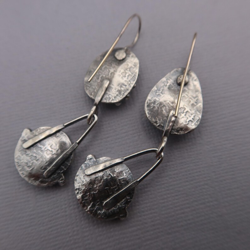 May include: A pair of silver-toned earrings with a textured, hammered finish. Each earring features an oval top piece connected to a circular bottom piece by a geometric design. The earrings have a hook closure.