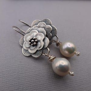 May include: A pair of silver earrings featuring flower-shaped pendants with detailed petal designs. Each earring has a dangling, lustrous white pearl. The earrings are designed with a hook closure.