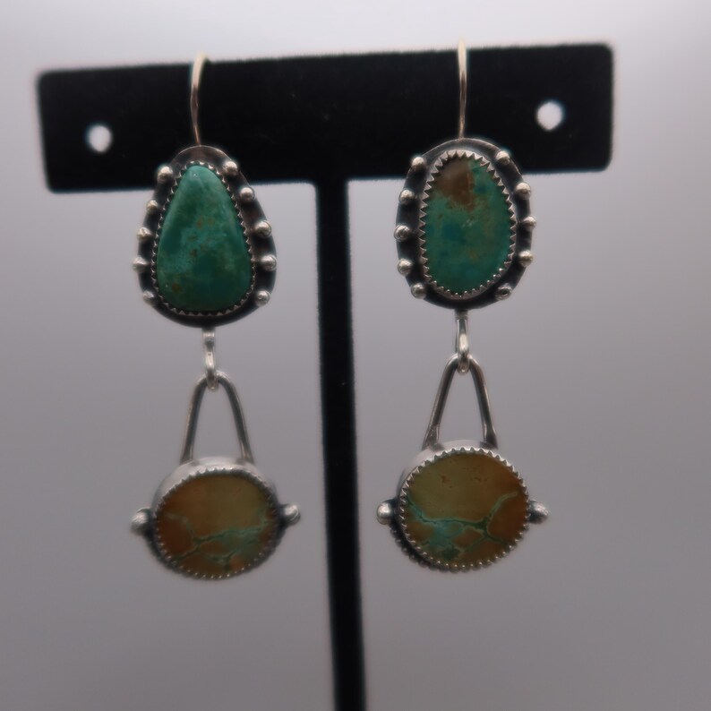 May include: Silver-toned dangle earrings featuring turquoise and tan-colored stones. The earrings have a teardrop-shaped turquoise stone at the top, a silver arch, and a circular tan stone at the bottom. The stones are set in a beaded silver frame.