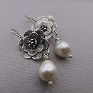 May include: A pair of silver-toned flower earrings with a pearl drop. The earrings feature detailed floral designs with a cluster of small beads at the center of each flower. The teardrop-shaped pearls are white and have a subtle luster.